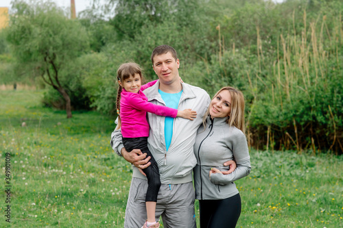 Sports family - mom, dad and daughter 5 years old in sportswear are hugging for a walk in the park