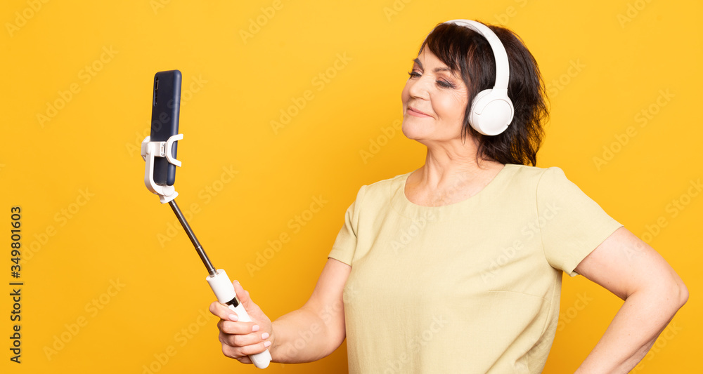 Beautiful Senior woman making video call by smartphone, monopod and wireless headphones over yellow background. Beautiful Senior woman making video call by smartphone, monopod and wireless headphones over yellow background.