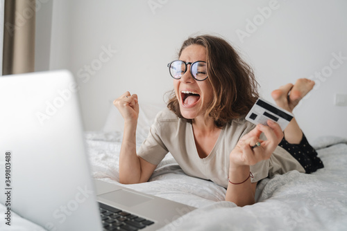 Pretty happy young curly woman with glasses spends time online shopping in bed, with a credit card in her hands with laptop