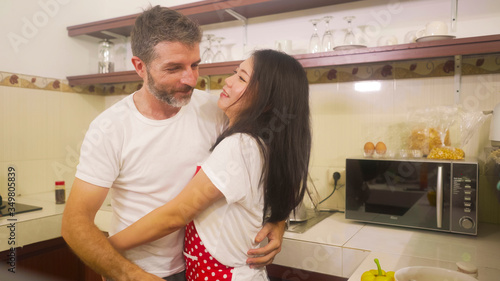 young beautiful and happy mixed ethnicity couple in love cooking together at home kitchen the woman Asian Chinese in red apron and her husband hugging her sweet