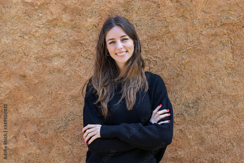 Pretty young brunette woman smiling looking at camera with her arms crossed standing next to a wall. She is wearing a black sweater and her hair is down. Vertical image. Pretty young brunette woman smiling looking at camera with her arms crossed standing next to a wall. She is wearing a black sweater and her hair is down. Vertical image.