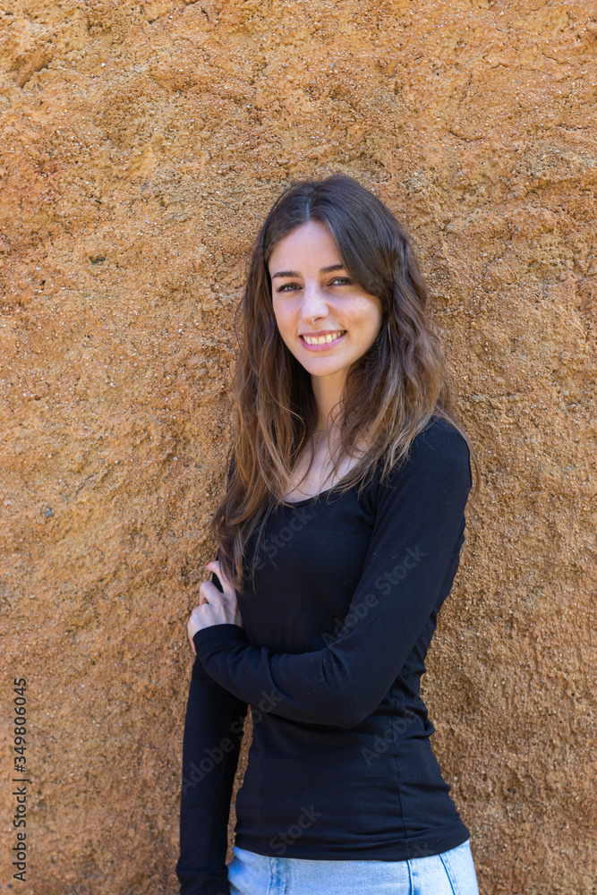 Pretty young brunette woman smiling looking at camera standing next to a wall. She is wearing a black T-shirt and her hair is down. Vertical image. Pretty young brunette woman smiling looking at camera standing next to a wall. She is wearing a black T-shirt and her hair is down. Vertical image.