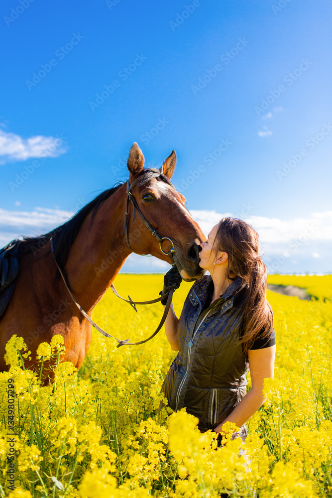 Fototapeta premium Young woman staying near brown horse in yellow rape or oilseed field with blue sky on background at sunny day. Horseback riding.