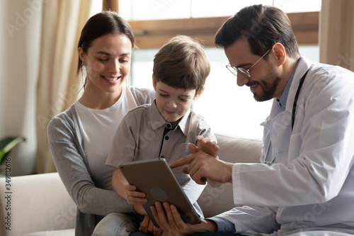 Positive male pediatrician sit on couch with little boy patient with mom show anamnesis on tablet, caring man doctor with small child and mom client use pad gadget for lab results in modern hospital