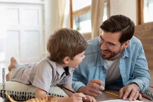 Happy loving young dad and little son lying on floor at home drawing in album together, smiling playful Caucasian father and small boy child have fun play painting on paper, enjoy family weekend