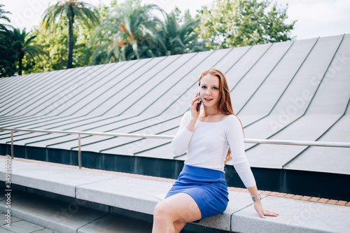 Portrait of charming caucasian female in casual wear sitting outdoors satisfied with mobile phone connection on conversation in roaming, pretty millennial woman looking at camera making cellular call