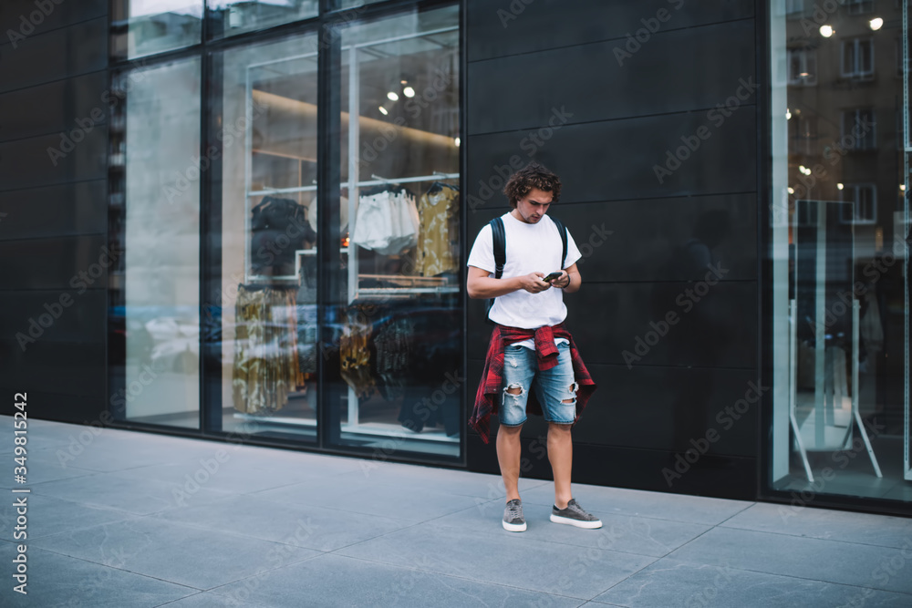 Pensive casually dressed caucasian traveler walking on city street using navigation app on mobile phone, concentrated young 20s checking notification about income message on smartphone on street