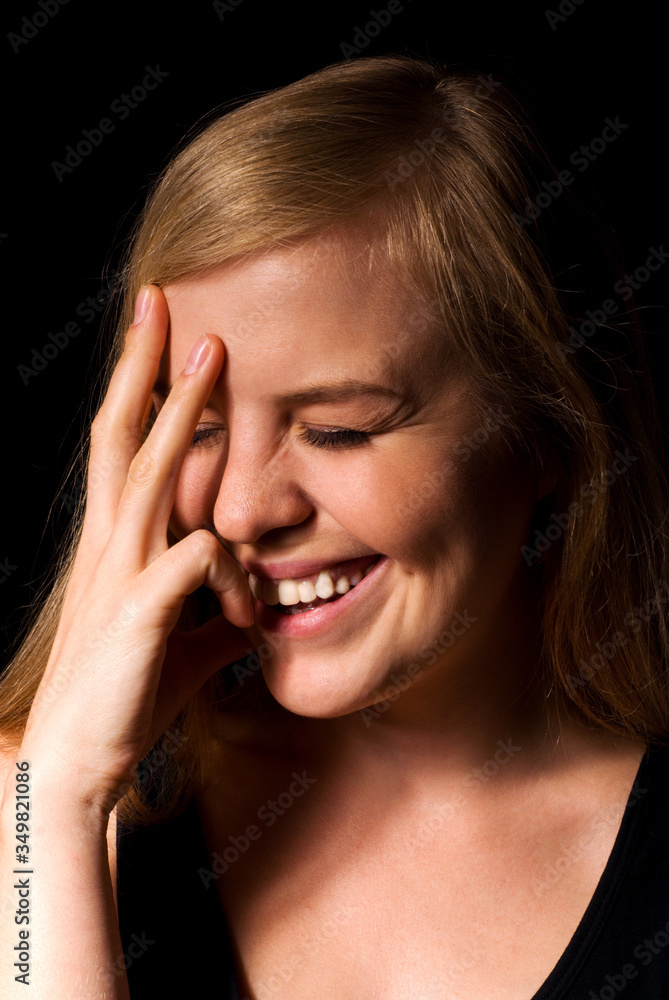 A young attractive blonde girl laughs sincerely, covering her face with her hand, a large portrait on a black background. A young attractive blonde girl laughs sincerely, covering her face with her hand, a large portrait on a black background.
