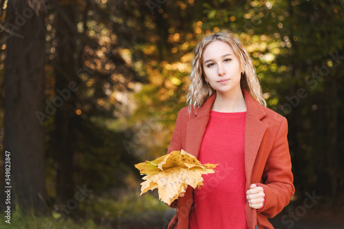 Portrait of an attractive young blonde in a red coat with a bouquet of yellow leaves in her hand. The concept of dreaming about forest walks during a virus pandemic. Autumn fashion in the forest