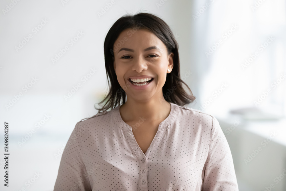 Head shot cheerful African woman with wide smile pose indoor. Mixed-race female blogger share positive news laughing look at web cam record video feels cheery, modern tech, happy girl portrait concept