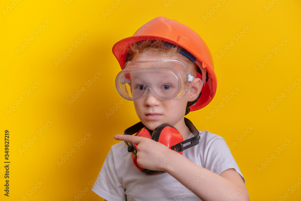 Cute boy in builder glasses, helmet and headphone to ear protection shown finger up over yellow background copy space Cute boy in builder glasses, helmet and headphone to ear protection shown finger up over yellow background copy space