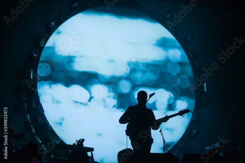 Silhouette of a guitarist playing against a large, moon-like backdrop on stage, creating a dramatic and surreal visual effect at a live music event.