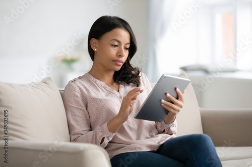 Focused beautiful African ethnicity woman sitting on couch in living room holding tablet gadget, freelancer working from home, housewife choose goods on-line surf websites stores, read news concept