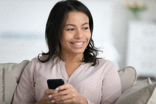 Beautiful african mixed-race young woman sit on sofa holds smartphone looking in distance, spend weekend web surfing chat with friends, using e-dating website services, enjoy new app wait call concept