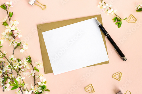 A flat lay composition with a mock-up of a white blank postcard for text, an envelope from craft paper, a pen, office clips and a branch of cherry with flowers on a pink background. Top view.