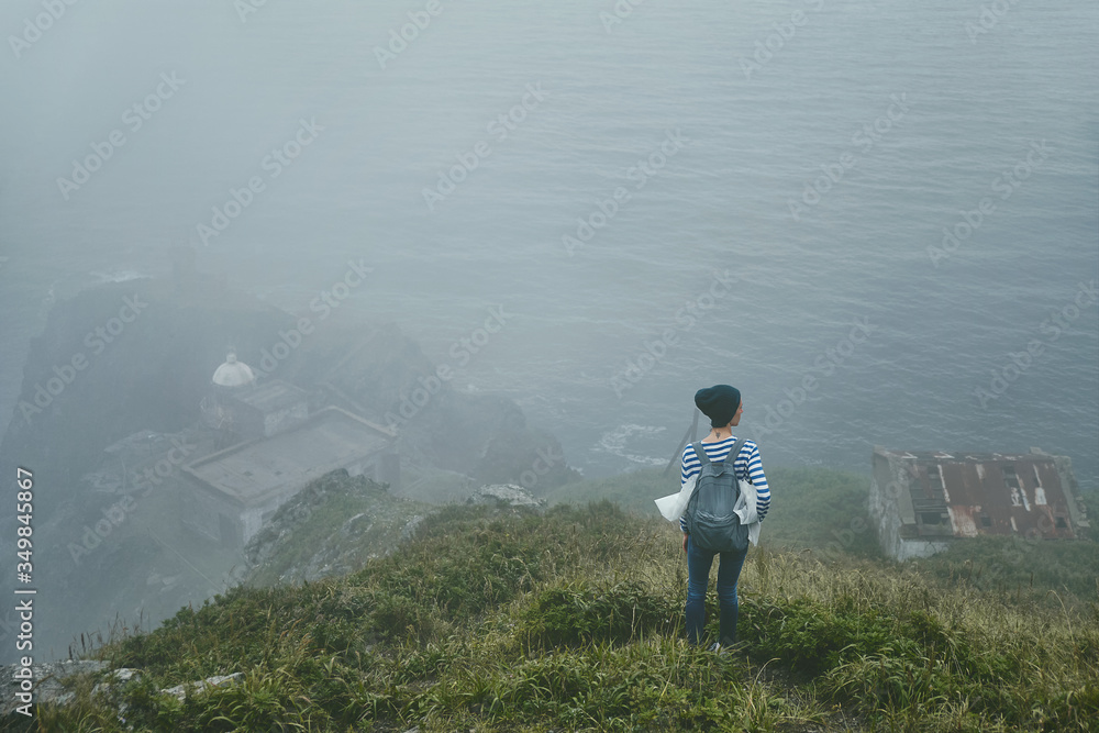 A young girl with a backpack and hat is standing on a steep cliff with a view of the old abandoned lighthouse and the sea on a summer foggy day. Tourism and outdoor concept.