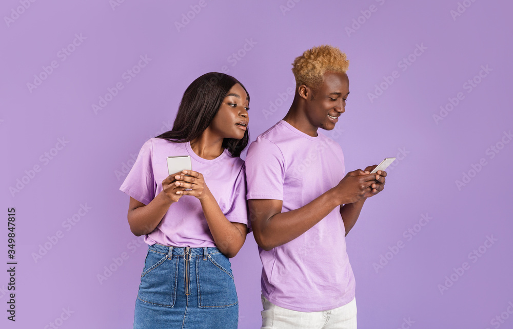Jealous black girl looking over her boyfriend's shoulder into his smartphone, watching his message on lilac background Jealous black girl looking over her boyfriend's shoulder into his smartphone, watching his message on lilac background