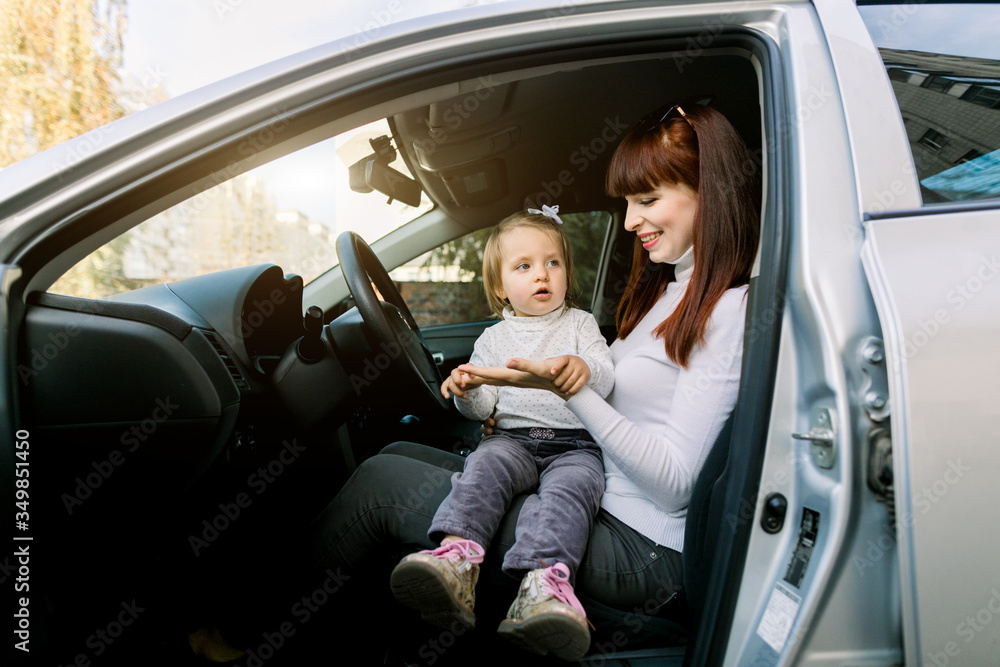 Pretty smiling young mother with a small child, cute baby girl, sits near the steering wheel of a car. Family trip, baby in a car Pretty smiling young mother with a small child, cute baby girl, sits near the steering wheel of a car. Family trip, baby in a car