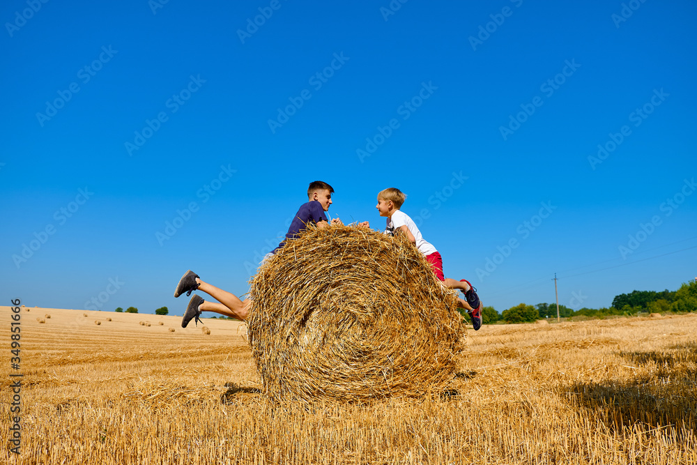 Teenagers in shorts and t-shirts jump on haystacks. Wheat hay on an agricultural field.  Children's day in nature. Friends having fun outdoors. Friends look each other in the eyes.