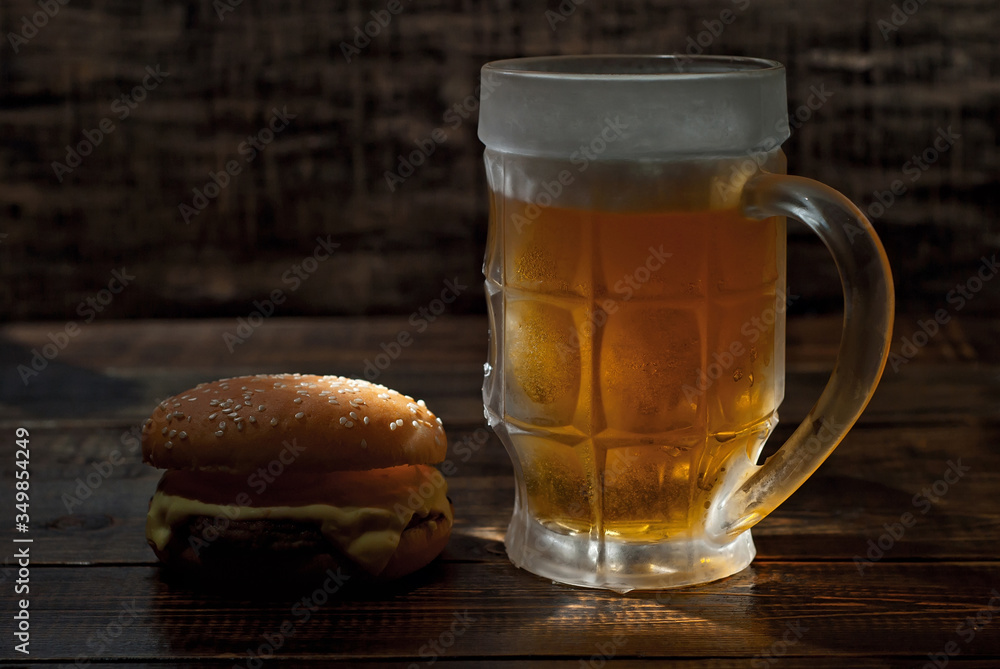 Cheeseburger with a glass of beer on a dark wooden table. Food on a black shabby background. Contrasting yellow light sparkles on the glass. Frozen mug with amber beer. Cheeseburger with a glass of beer on a dark wooden table. Food on a black shabby background. Contrasting yellow light sparkles on the glass. Frozen mug with amber beer.