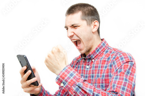 Young man in a plaid shirt with a smartphone on a white background. The guy holds the smartphone in front of him and screams, clenching his fist in front of him with joy. Man rejoices, screaming, win