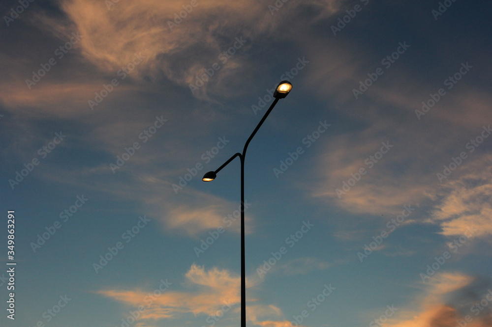 street lighting a lamp post with two lights against a cloudy sky is illuminated by the setting sun