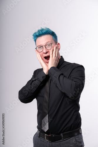 Young stylish european man with blue hair expressing amazement, shock and excitement, opening his mouth widely and touching his face