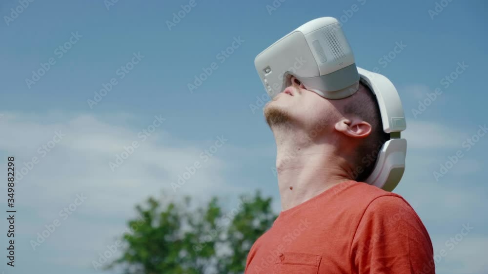 Man in a virtual reality helmet raises his head up. The guy in the park in a red T-shirt on a background of blue sky and green trees looks VR glasses. 4K footage Man in a virtual reality helmet raises his head up. The guy in the park in a red T-shirt on a background of blue sky and green trees looks VR glasses. 4K footage