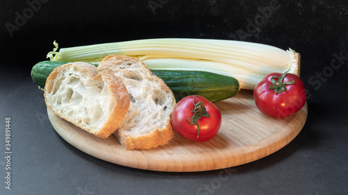 Two slices of white beautiful airy white bread, fresh vegetables - cucumbers, tomatoes and celery, all on a round wooden cutting board on a black background close-up.