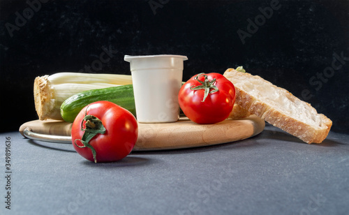 Slices of white beautiful airy white bread, yogurt, fresh vegetables - cucumbers, tomatoes and celery, all on a round wooden cutting board on a black background close-up, side view. Place for text.