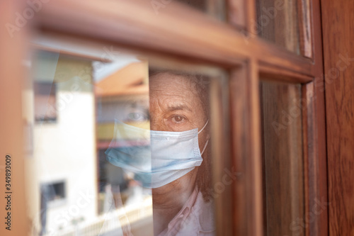 Ritratto di anziana che guarda dalla finestra di casa sua con l mascherina e aria triste photo