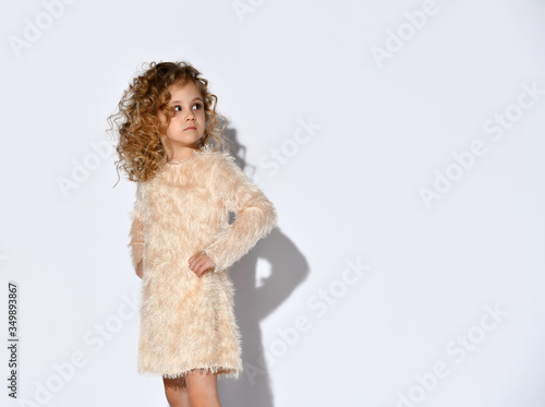Little blonde curly kid in fluffy dress and shoes. She has put her hands on hips, posing standing sideways isolated on white
