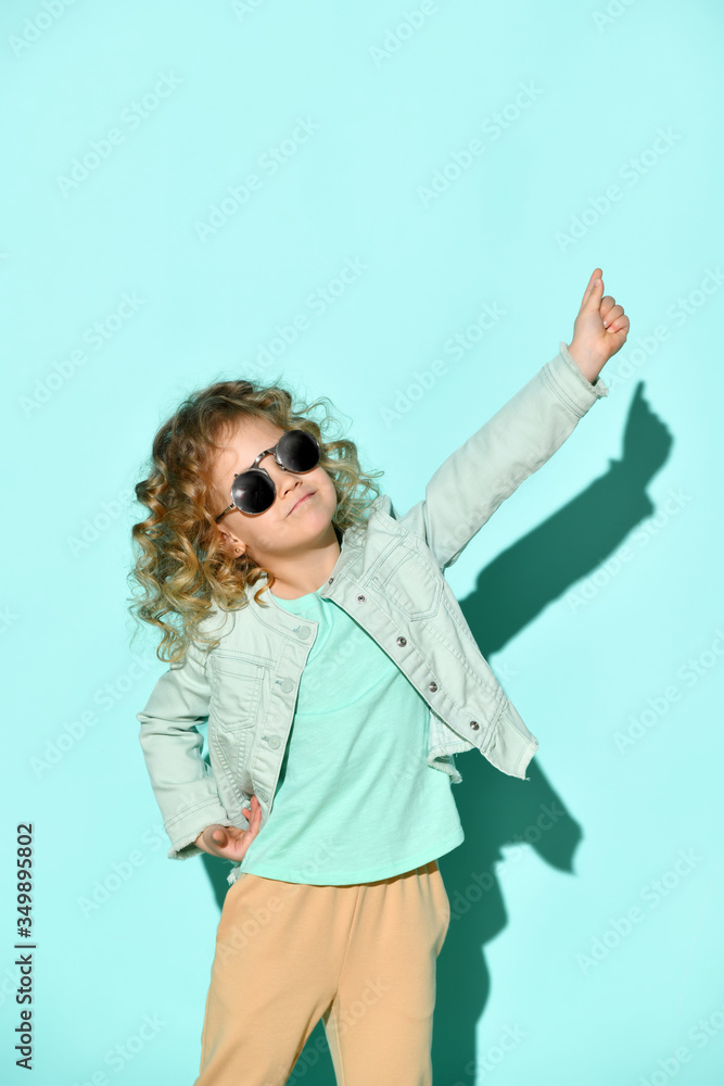 Cute little curly girl with black non-transparent glasses on face stands raising one hand pointing two fingers up. Studio shot isolated on turquoise Cute little curly girl with black non-transparent glasses on face stands raising one hand pointing two fingers up. Studio shot isolated on turquoise
