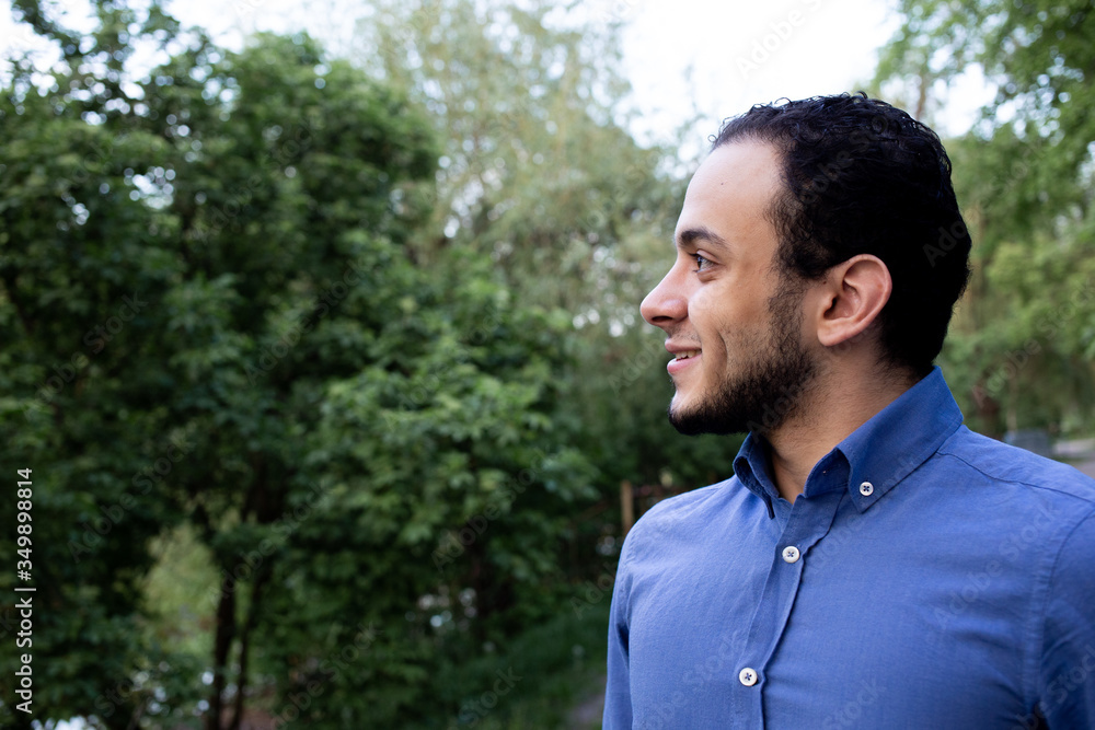 Face closeup profile of the young man from Middle Eastern. Closeup portrait of a smiling man from Egypt in blue shirt in the park. Trees on the background. Face closeup profile of the young man from Middle Eastern. Closeup portrait of a smiling man from Egypt in blue shirt in the park. Trees on the background.