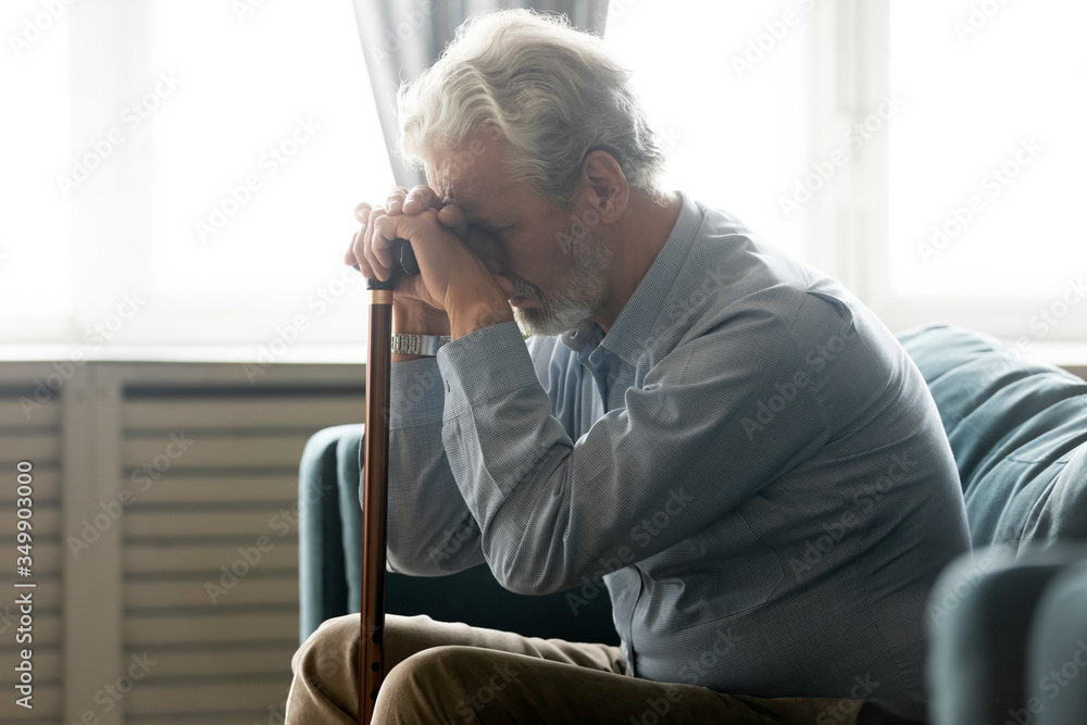Depressed mature old retired man sitting on sofa, holding wooden stick cane in hands, feeling stressed alone at home. Disabled middle aged senior grandfather suffering from loneliness in living room. Depressed mature old retired man sitting on sofa, holding wooden stick cane in hands, feeling stressed alone at home. Disabled middle aged senior grandfather suffering from loneliness in living room.
