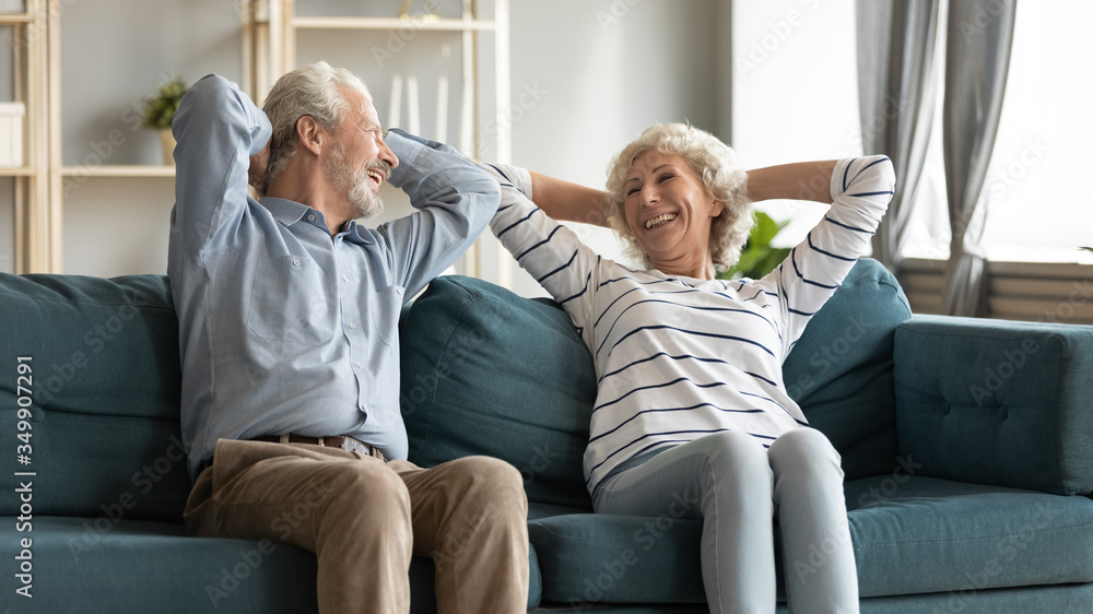 Happy middle aged retired married couple resting on comfortable sofa with folded hands behind hand. Relaxed pleased senior mature man enjoying free leisure lazy weekend time at home with older wife. Happy middle aged retired married couple resting on comfortable sofa with folded hands behind hand. Relaxed pleased senior mature man enjoying free leisure lazy weekend time at home with older wife.