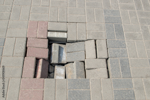 Fotografi Walkway paved with gray and pink blocks with break in sidewalk, can see sunken earth and several tiles that have fallen into the hole