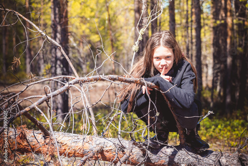 Beautiful girl in warm sweater sits and shows finger at her mouth about silence in pine forest.