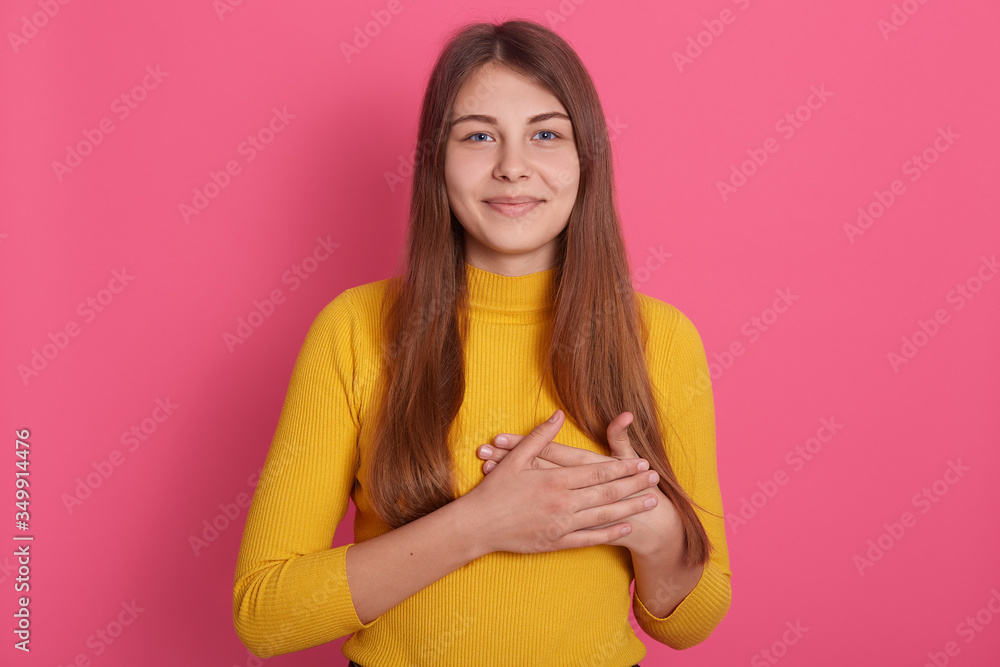 Obraz premium Studio shot of young Caucasian lady posing indoor against pink background, keeping hands on chest, has grateful facial expression, wearing casual attire, looking at camera, being thankful to someone.