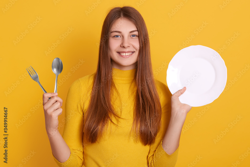 Portrait beautiful young caucasian woman wearing casual shirt, standing with clean plate, spoon and fork, posing isolated over yellow background, looking directly at camera with charming smile. Portrait beautiful young caucasian woman wearing casual shirt, standing with clean plate, spoon and fork, posing isolated over yellow background, looking directly at camera with charming smile.