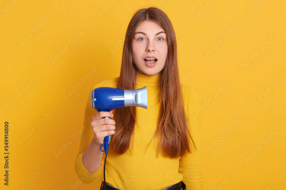 Horizontal shot of beautiful young woman holding blue hairdryer in hand, posing isolated over yellow studio background, having astonished facial expression,looks surprised, poses with opened mouth. Horizontal shot of beautiful young woman holding blue hairdryer in hand, posing isolated over yellow studio background, having astonished facial expression,looks surprised, poses with opened mouth.