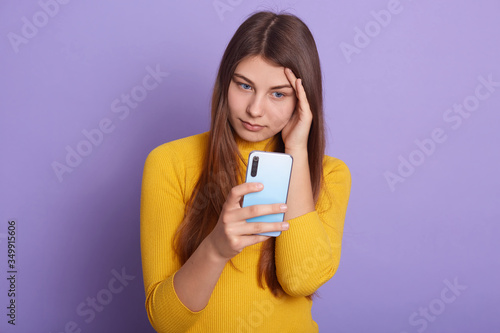 Valokuva Closeup portrait of sad female dresses casual outfit, keeping hand on her head, holding smart phone in hands, having troubles, posing isolated over lilac studio background