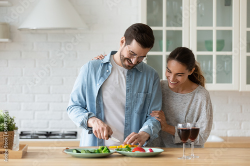 Happy millennial husband and wife stand at counter cooking dinner salad together, smiling young Caucasian couple have fun preparing food in home kitchen enjoy family weekend or celebration