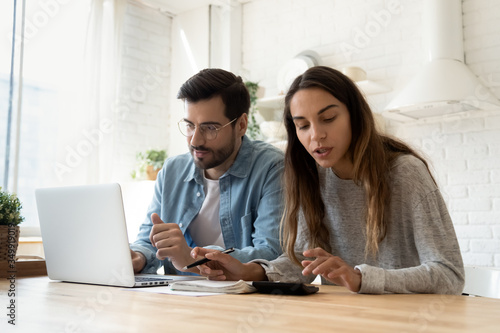 Concentrated young couple sit at desk in kitchen pay bills on computer online calculate finances together, millennial husband and wife manage household expenses use Internet banking system on laptop