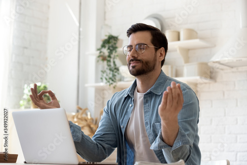 Calm millennial Caucasian man in glasses sit at table distracted from computer work meditate in kitchen, peaceful young male practice yoga relieve negative emotions at workplace, stress free concept