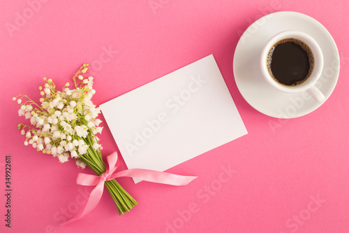 Blank white sheet of paper, black coffee in the cup and bouquet of lilies of the valley on the pink background. Top view. Copy space.