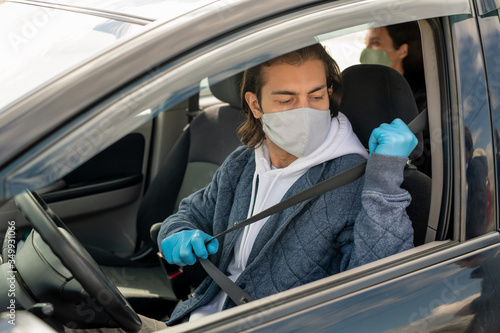 Young man in gloves and mask going to put seatbelt while sitting in car
