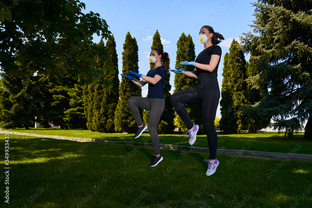 Obraz premium Two girls do sports exercises in protective masks in the open air in park. Social distancing. Alternative fitness workouts during epidemic and quarantine. Covid 19 pandemic. Coronavirus