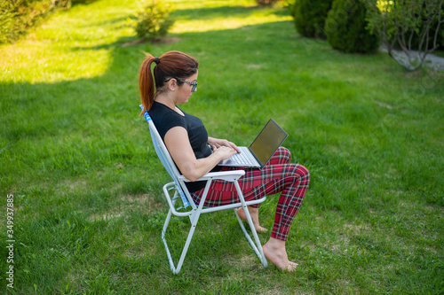 A woman with glasses sits in a folding chair and types on a laptop. Female freelancer remotely works in the garden of the cottage during quarantine. Work is a pleasure. © Михаил Решетников