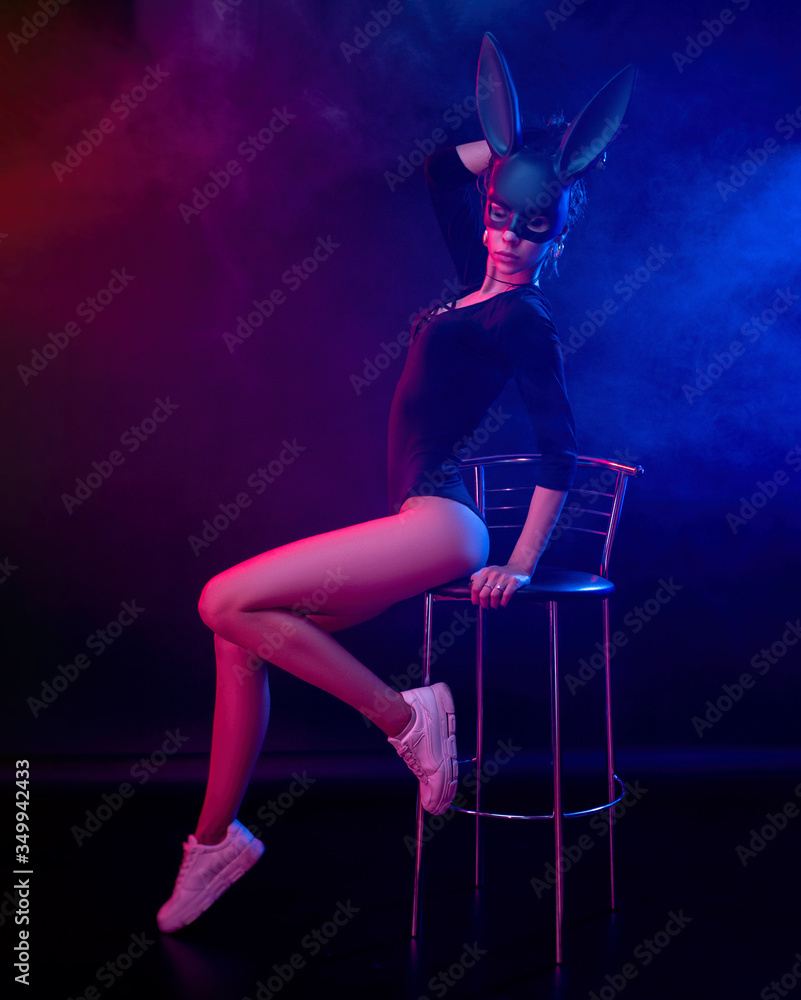 a beautiful girl in a bodysuit poses in a photo Studio on a dark background in neon light. On the face of a rabbit mask a beautiful girl in a bodysuit poses in a photo Studio on a dark background in neon light. On the face of a rabbit mask